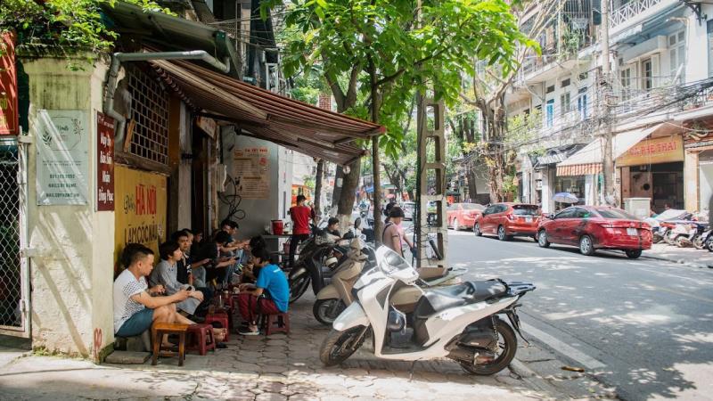 Một quán cà phê vỉa hè dưới bóng cây, nắng rọi qua từng ô cửa